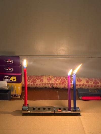 Three candles burn brightly in a silver-colored metal menorah. The candle colors are red, pink, and blue, from bottom to top. Boxes of matches and a lighter sit underneath. The menorah stands in front of a cardboard surface next to a cushion with a red and cream pattern. A light gray painted wall is visible on the left.