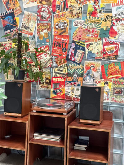 The image depicts a corner of a room, likely a living space or a small shop, with a vintage aesthetic. The dominant feature is a wall adorned with a collage of retro-themed posters, primarily featuring advertisements and movie posters from the mid-20th century. These posters display graphics of old cars, movie stars, food and drink advertisements, and travel scenes, all rendered in a stylized, colorful, and somewhat kitschy manner. The posters are a mix of English and French text.
In the foreground, two wooden speaker cabinets are placed on modular wooden shelving units. The speaker on the left is black with a wooden trim, and a Klipsch logo is visible. On top of the speaker, a green potted plant with large leaves is situated on a saucer. Next to it, a vintage turntable with a vinyl record is placed on the shelf. The speaker on the right is also black with a wooden trim and a Klipsch logo. Below it, the shelves contain stacked vinyl records and a small stack of books.
The wall behind the posters appears to be made of translucent glass blocks, allowing some light to filter through, creating a diffused background. A goose-neck desk lamp with a silver metallic finish is positioned between the two speaker setups, illuminating the turntable. The overall color palette is warm, with rich browns from the wood, vibrant colors from the posters, and the green of the plant. The lighting suggests an indoor setting, possibly in the late afternoon or evening, with artificial light from the lamp.
There are no people visible in the image. The scene is static and appears to be a curated display, emphasizing a retro or nostalgic theme. The context provided regarding Tam Kỳ, Vietnam, is not directly evident from the visual elements of the image itself.