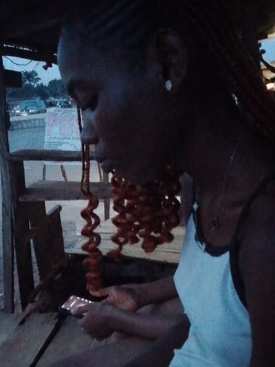 The image depicts a dark-skinned woman with braided hair, some of which are styled into red, curly tendrils that hang down. She is wearing a light-colored tank top and a necklace. Her head is tilted downwards as she looks at a smartphone in her hands. She appears to be seated in a rustic outdoor space, possibly a covered porch or simple structure with wooden supports and a mesh-covered opening, suggesting a location in or around Urualla, Nigeria, given the prompt. In the background, some trees and a road with parked vehicles are visible. The lighting in the image is subdued, with shadows obscuring details.