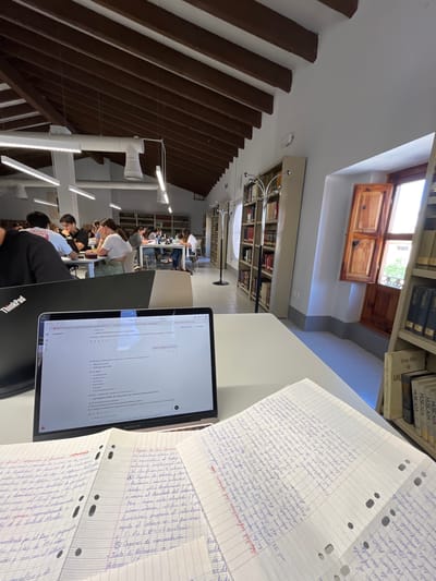 The image shows a well-lit study area in a library in Torrent, Spain. The view is from a student's perspective at a table with notes and a laptop. Several other students are studying at the tables. Bookshelves line the walls, and there are skylights.