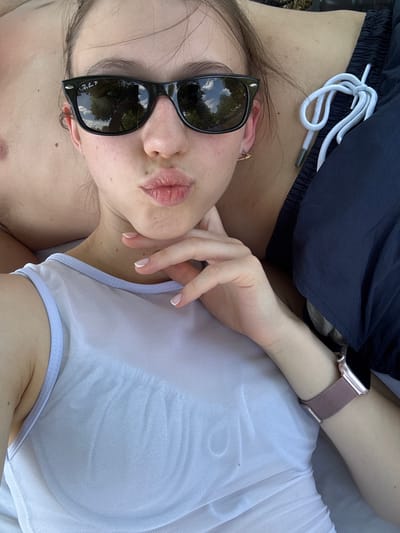 A woman is lying down and pouting with her lips pursed. She is wearing sunglasses and a light-colored top. She is also wearing a watch on her wrist. The background is blurred, but appears to be outdoors. There is no specific information about the location in Bürgstadt, Germany.