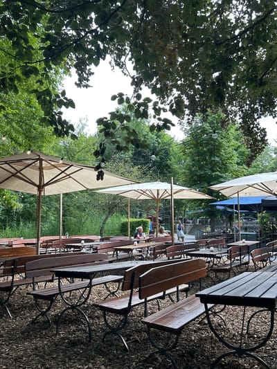 The image depicts an outdoor seating area with numerous wooden tables and benches arranged on a ground covered with wood chips. Large, light-colored umbrellas provide shade over some of the tables, and string lights are visible hanging from branches and poles. The setting appears to be a beer garden or restaurant with abundant greenery, including lush trees and bushes surrounding the area. In the background, several people are seated at tables, some appearing to be conversing or dining. The overall atmosphere suggests a relaxed, outdoor dining or gathering space. While the image does not explicitly show any landmarks or signage to pinpoint a specific location in Heilbronn, Germany, the style of the seating and the presence of umbrellas are common features of German beer gardens.