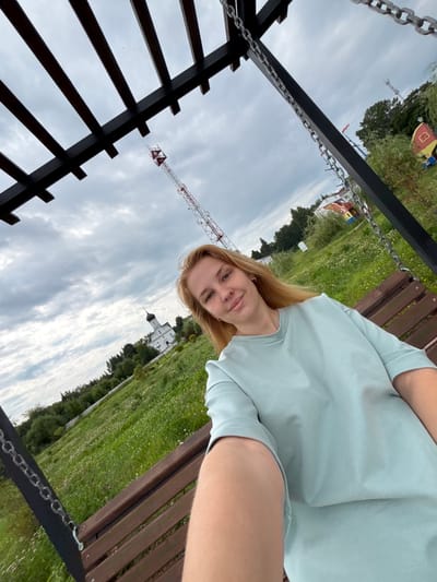 A young woman with blonde hair takes a selfie while sitting on a wooden swing in what appears to be a park or outdoor recreational area. She is wearing a light blue t-shirt. In the background, a white church with a dark dome is visible, surrounded by green trees and a field of grass with small white flowers. A radio tower is also present in the distance, along with other structures that suggest a town or amusement park. The sky is overcast. This scene is located in the city of Breytovo, Russia.
