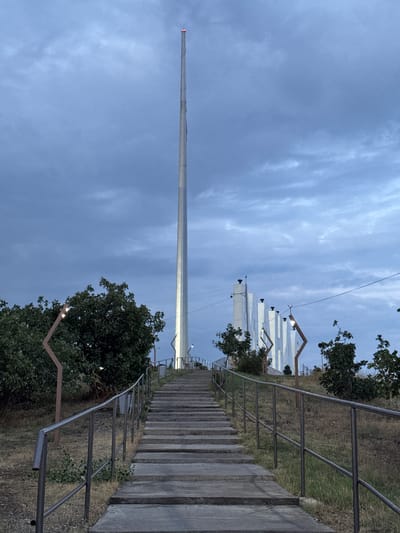 The image depicts a tall, slender metallic flagpole topped with red lights, situated in the city of Manas, Kyrgyzstan. A wide concrete staircase with metal railings leads upwards towards the flagpole. The surroundings are characterized by dry grass, sparse trees, and modern-looking white structures in the background. The sky is overcast with a palette of blue and grey clouds, indicating either dawn or dusk. There are no people visible in the scene.