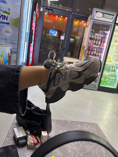 The image is a close-up shot of a person's leg and foot, wearing a New Balance sneaker. The foot is propped up on a table, and a black handbag is visible below it. On the table, there are several small items, including a pack of "TEREA" cigarettes, a wrapper for what appears to be a candy bar, and a pink and white box. In the background, there is a storefront with refrigerated display cases stocked with beverages. The overall setting suggests a convenience store or small shop. The location is identified as Satovcha, Bulgaria.