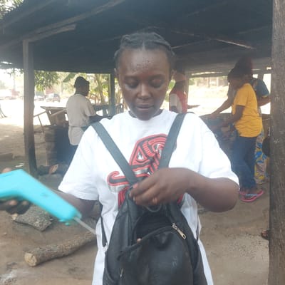 The image captures an outdoor food preparation or market scene in Assai Sabongida, Nigeria, under a dark, low-lying canopy structure. In the foreground, a dark-skinned female with braided hair, wearing a white t-shirt with a red and black graphic and a black backpack, is seen looking downwards. She holds a light-blue, pistol-grip device with a metallic probe in her left hand, while her right hand is positioned on the top zipper or flap of her backpack. The immediate ground is dirt, with two cut logs visible. In the mid-ground, beneath the canopy, several individuals are actively engaged in cooking or tending to food. To the left, a male in a light-colored shirt stands near a stone cooking structure. To the right, two females, one in a yellow t-shirt and another in a patterned dress, are interacting with additional cooking or preparation stations. A dirt road, distant vehicles, and vegetation are discernible in the background beyond the covered area.