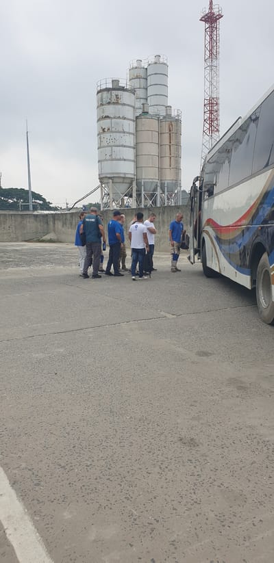 In Rooppur, Bangladesh, a group of approximately ten individuals, predominantly men, are gathered in an outdoor industrial area. The setting features several tall, cylindrical industrial silos in the background, some appearing weathered and rusted. A red and white transmission tower is visible behind the silos. In the foreground, a portion of a bus with a distinctive colorful stripe design is parked. The individuals are dressed in various casual attire, including t-shirts, pants, and one person in a dark green uniform with text that appears to read "POWKA ARCHIVER." Some individuals are standing and conversing, while others appear to be waiting. The ground is paved with concrete, showing some cracks and stains. The sky is overcast.
