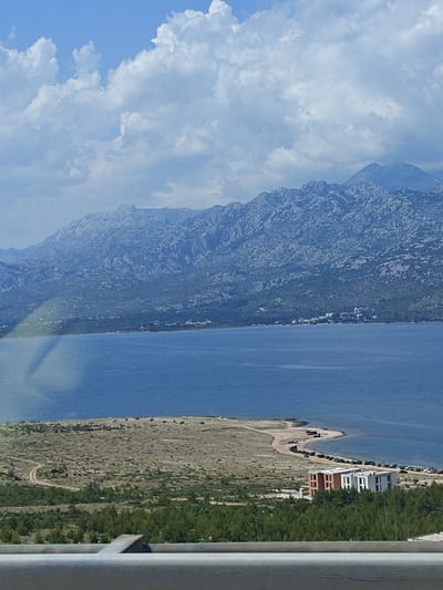 A landscape image captures the Adriatic Sea and a mountainous backdrop in Rovanjska, Croatia. The foreground is dominated by a concrete barrier and a dense green forest. Beyond the forest, a sandy peninsula juts into the bright blue sea, with a dirt road winding its way across it. On the peninsula, a modern building complex with white and orange facades stands, surrounded by parked cars. In the distance, a small cluster of houses is visible along the coast, nestled at the foot of a rugged, rocky mountain range. The sky above is a vibrant blue, dotted with fluffy white clouds.
