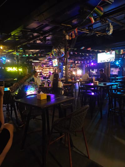 The image depicts a vibrant, dimly lit bar or restaurant scene, likely in Ban Klong Haeng, Thailand, at night. The setting is characterized by an abundance of colorful flags from various nations hanging from the ceiling and walls, suggesting an international theme. Disco balls and neon lights cast a festive and energetic ambiance.

Several people are visible, though their features are often obscured by the low lighting and the angle of the photograph, which appears to be taken from a slightly elevated perspective and rotated. In the foreground, a person's arm and shoulder are visible, suggesting the photographer is at a table.

There are multiple tables and chairs arranged throughout the space. One table in the middle ground has two individuals seated, one with their back to the camera and another facing away. A screen displaying what looks like a sporting event or a video is mounted on the wall to the left. Behind the bar area, shelves are stocked with bottles, typical of a bar.

The overall mood is lively and social, consistent with a popular nighttime entertainment venue. The presence of multiple flags and the busy atmosphere hint at a tourist-friendly location. The lighting is predominantly artificial, with blues, purples, and yellows creating a dynamic visual experience.
