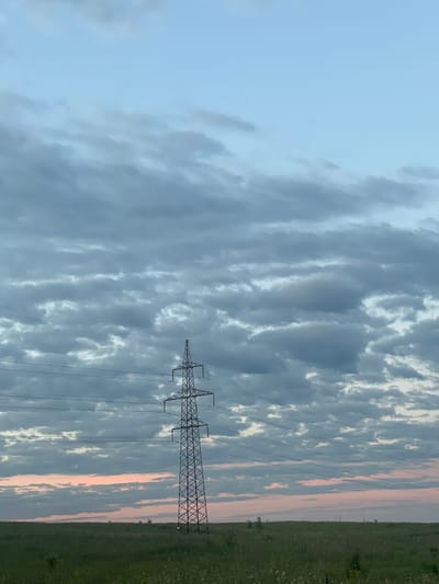 A power line tower stands in a field of green grass under a cloudy sky. The sky is a mix of blue and gray clouds, with a hint of pink near the horizon. The power line tower is tall and made of metal, with wires stretching across the frame. The field is flat and appears to be undeveloped. There are no people or animals visible in the image. The scene is calm and serene, with a sense of vastness and openness. The location is the city of Stroykeramika, Russia.