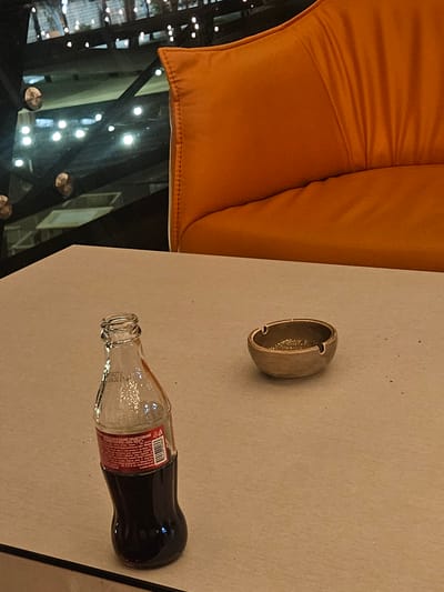 The image depicts an overhead view of a table with a few objects on it, set against a backdrop of an orange upholstered cushion. The table surface is a light, neutral color.

On the table, there are two primary objects. One is a small, shallow ashtray, appearing to be made of a metallic material with a subtle sheen. The other object is a glass bottle of Coca-Cola, lying horizontally on its side. The bottle is partially filled with the dark liquid, and its label, which is red and white, is visible. The bottle's neck and cap are also visible.

The orange cushion appears to be part of a seating arrangement, possibly a couch or booth. To the left of the table, the background shows a reflective surface, likely a glass table top or window, revealing a blurry view of lights and possibly indoor structures. These lights suggest an indoor setting, possibly at night or in a dimly lit environment.

The scene appears to be set in a casual or dining environment. There are no people visible in the image. The overall mood is still and uneventful, focusing on the arrangement of the objects on the table. The lighting is subdued, casting soft shadows. The reflection in the background provides some context of a place with ambient lighting. The visible text on the Coca-Cola bottle includes "Coca-Cola" and other product information in smaller print, likely in English or a Slavic language.
