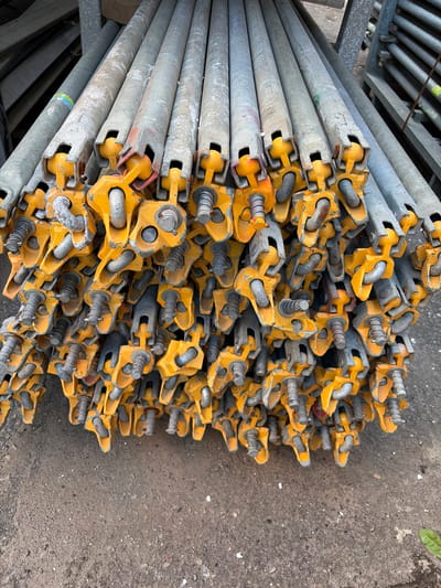 A close-up, top-down shot shows a large stack of metal scaffolding poles. The poles are cylindrical and appear to be made of galvanized steel, with some visible scuff marks and rust. Each pole has a bright yellow, metal attachment at one end, with a U-shaped hook and a threaded bolt with a spring mechanism. These attachments are designed for connecting scaffolding sections. The poles are stacked horizontally and diagonally, filling most of the frame.
The background is a coarse, grey concrete surface, possibly a construction site or industrial area. Some small white debris is scattered on the concrete. In the upper left corner, a glimpse of a metal rack holding more poles or similar equipment is visible. The lighting suggests it is daytime, and the overall scene conveys a sense of industrial material storage. There are no people or animals visible in the image. No text is legible on the scaffolding poles or in the surrounding environment. The location context is Syktyvkar, Russia.