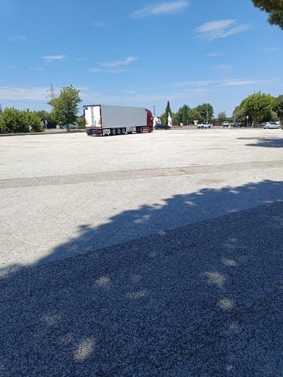 An empty, sunlit parking lot with a large truck is pictured under a bright blue sky with scattered clouds. The truck, a semi-trailer with a grey trailer and a red cab, is parked in the middle distance on the right side of the frame. The parking lot pavement is light grey and cracked, with some darker patches and a prominent shadow of trees cast across it from the left. In the background, there are more trees and a few other vehicles parked, including a white SUV. To the left of the truck, a white statue is visible. The scene appears to be in an open area with minimal activity.