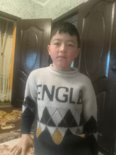 The image displays a young male, appearing to be a child, standing in a frontal pose within an indoor environment located in the city of Pakhtakor, Uzbekistan. He has short, dark hair and a light complexion, with visibly flushed cheeks. His expression is neutral as he looks directly into the camera. He is wearing a long-sleeved, high-neck sweater that is primarily light gray. The upper chest area features the partial word "ENGLA" in dark gray lettering. Below this, the sweater transitions into a pattern of dark gray, light gray, and mustard yellow argyle diamonds, continuing down the sleeves. The individual's right hand is visible in the lower-left foreground, resting on an unidentifiable light-colored surface. The background consists of two dark brown wooden doors, possibly paneled, positioned behind the subject. To the left of the left door, a vertical section of yellowish fabric or curtain is visible, partially obscuring a sheer white curtain. The floor shows a patterned rug in earth tones with abstract or floral motifs. The walls and ceiling are light-colored, showing minor textural inconsistencies. The scene is illuminated by ambient indoor light, resulting in a slightly grainy image.