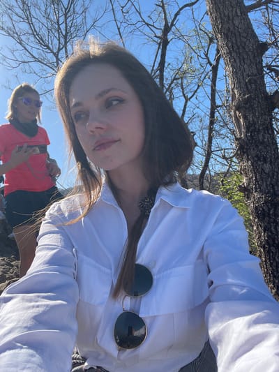 A daytime, medium-shot, outdoor photograph depicts a young woman wearing a white collared shirt with gold-rimmed sunglasses clipped to the collar. The woman has long brown hair. Behind and below her is another person, perhaps a woman, with sunglasses and a pink shirt. In the background is a tree and blue sky with branches. This could be Bled, Slovenia due to the environmental features.