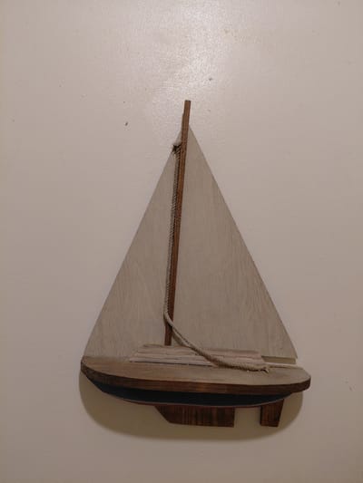 A wooden boat decoration hangs on a light-colored wall. The boat is a simplified design, featuring a triangular sail and a multi-layered hull. A rope runs along the top of the sail. The environment appears to be an interior space with a plain wall surface. There is no evidence to suggest the location of Cutler Bay, United States of America. There are no people or actions present.
