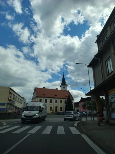 The image depicts a street scene in Bernartice, Czechia, on a cloudy day with a blue sky visible. In the foreground, a white van with the text "DOPRAVNÍ ZNAČENÍ JÁNEV" is driving across a pedestrian crossing. To the left, a building with a "Supermarket" sign is visible. In the background, a yellow building with a red-tiled roof and a prominent church tower with a clock is situated. To the right, a dark-colored building with windows and a street light stands beside a sidewalk where a person on a scooter is present. Several other cars are parked or driving on the street. The overall impression is of a typical town square with a mix of commercial and religious architecture.