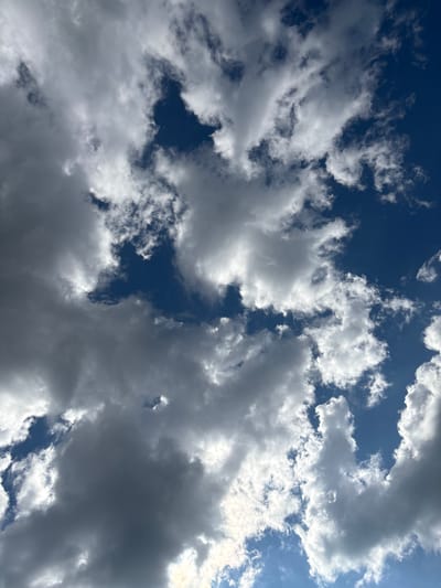 A high-angle shot captures a dramatic sky filled with a mix of dark, heavy clouds and bright, fluffy white clouds against a deep blue sky. Sunlight is breaking through in several areas, illuminating the edges of the white clouds and creating stark contrasts. The image appears to be taken from a ground-level perspective, looking directly upwards. There are no people, structures, or any other discernible elements of the city of Kzyl-Bairak, Russia, visible in this image; it solely depicts the sky.