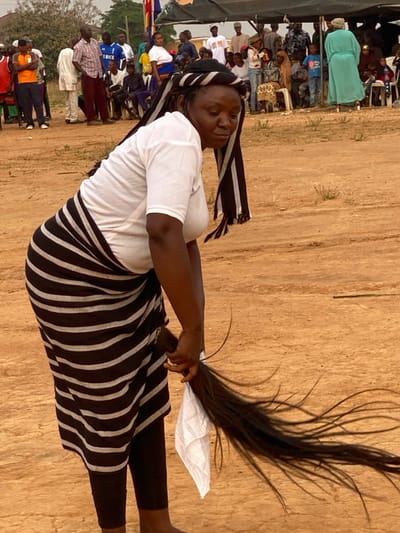 A woman stands outdoors on a dirt ground, a crowd of people visible behind her. She wears a white t-shirt, a striped black and white skirt, and black leggings. A black and white striped scarf adorns her head. She holds a long, dark ponytail in her hand, secured with a white cloth. The background is a field of dirt with some greenery visible in the distance. The photograph is taken at an angle.