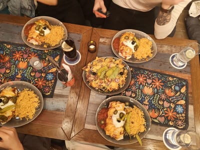 The image displays an overhead view of a dining table within a restaurant in Aschaffenburg, Germany. The specific address or name of the establishment within Aschaffenburg is not identifiable from the visual content.
The table surface is composed of distressed wooden planks exhibiting varying tones, including grey, brown, and light blue. Two dark fabric placemats, patterned with vibrant floral and foliage designs in colors such as orange, blue, green, and pink, are laid out.
Five plates of food are arranged on the table. Four plates each contain two cheese-covered rolled items, consistent with enchiladas, alongside a mound of yellow rice, a portion of red salsa, a dollop of white cream, sliced green jalapeños, black olives, and whole pickled green peppers. A fifth plate holds a large serving of nachos, topped with melted cheese, red onion rings, black olives, green jalapeños, and a substantial quantity of guacamole.
Beverages include two glasses with a dark liquid and a lemon slice, placed on blue and white coasters, and two clear glasses of water, each with a lemon slice, also on similar coasters. Two small, decorative votive candle holders, each containing a lit candle, are positioned on the table, emitting a warm light. A container filled with assorted cutlery and a napkin dispenser are also present.
Portions of at least four individuals are visible around the table, indicating a group dining. One person in the upper left wears a blue denim jacket. Another individual in the upper center, wearing black pants and a white t-shirt, holds a red-cased smartphone and exhibits a visible tattoo on their forearm. Hands and arms of other diners are discernible in the lower left and lower right sections of the frame. The scene captures a social dining event.