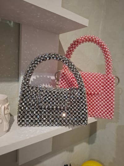 Two beaded handbags, one silver and one pink, are displayed on a white shelf. The silver handbag is in the foreground and is covered in faceted, reflective beads. The pink handbag is behind and to the right of the silver one, and is made of smooth, spherical pink beads. The handbags are illuminated by light that casts dappled shadows on the white shelf and the textured wall behind them. A white decorative object is partially visible on the left side of the shelf. The scene is indoors, likely in a retail or display setting. No people are present in the image. The location is the city of Korenovsk, Russia.