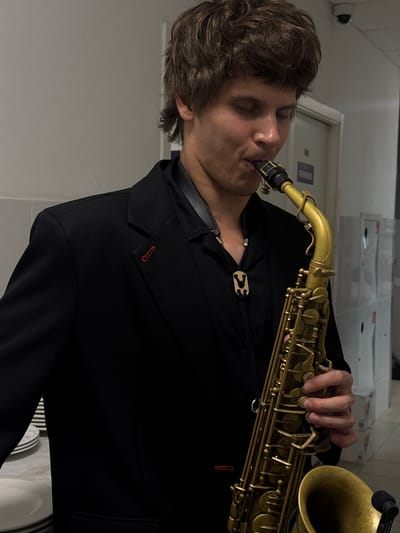 A young man with tousled brown hair is captured in a medium shot, playing a golden saxophone. He wears a black suit jacket over a black shirt, accessorized with a bolo tie featuring a unique decorative clasp. His eyes are closed as he focuses on his music. The background appears to be an indoor, possibly backstage or service area, with a wall of grey and some white lockers or cabinets. Stacks of white plates are visible on a surface to the left of the frame. The lighting is dim, suggesting an indoor event setting. There are no discernible landmarks that would place the scene specifically in the city of Истра, Russia.