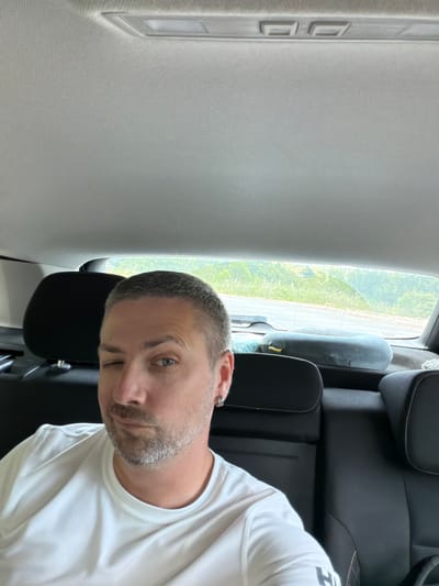 A man with short grey hair and stubble is sitting in the back seat of a car. He is wearing a white t-shirt and winking at the camera. He has a small hoop earring in his left ear. The car's interior is visible, including black seats and the headliner. Outside the car, through the rear window, a blurred landscape of green trees and foliage can be seen, suggesting a rural or suburban environment. The image appears to be taken in Svinjišta, Montenegro.