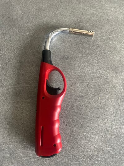 A single red utility lighter is depicted resting on a textured, light-grey surface in Oberhausbergen, France. The lighter features a red plastic handle with a distinct pistol-grip design, including finger grooves. A black trigger mechanism is located within a circular guard. Extending from the top of the handle is a flexible, corrugated metallic neck, which bends in an arc towards the upper right of the frame. This neck terminates in a rigid, silver-colored metallic nozzle with three rectangular apertures visible on its upper surface. The underlying grey surface displays numerous fine, linear scratches and scattered specks across its entirety. No people, actions, or interactions are visible.