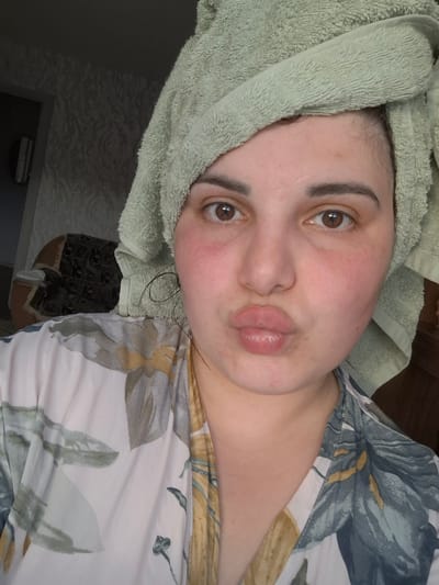 A person is lying down, with a green towel wrapped around their head. They have fair skin, brown eyes, and are making a kissing face. The person is wearing a floral print top. The location appears to be indoors, with a patterned wall and furniture visible in the background. There is no indication this is in the city of Амзя, Russia.