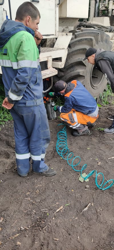 The image shows three individuals in work attire near a large vehicle, possibly a tractor or heavy machinery, in an outdoor setting, likely in Kovalivs'ke, Ukraine.
One individual is lying down, dressed in blue and green work overalls, with their hands near their face. A second individual is bending down, wearing a blue jacket over an orange safety vest and a cap, appearing to work on a component near the vehicle's wheel. A third person in dark clothing and a cap is standing near the wheel, looking down.
A coiled, light blue hose is visible on the ground near the individual who is bending over. The ground is dirt, with some vegetation visible. The vehicle has a large, weathered tire and a white body.
