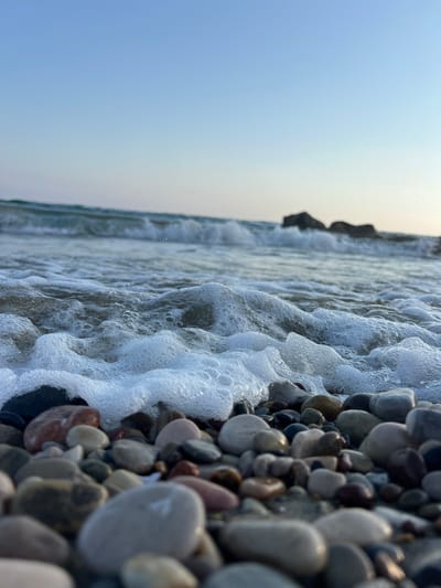 The media file displays a close-up, low-angle shot of a pebble beach with waves gently washing ashore. The pebbles in the foreground are varied in size and color, ranging from light grey and white to darker shades of brown and black, with some appearing wet and shiny from the water. The water itself is foamy and white as it recedes from the shore, creating interesting patterns and textures. In the background, the ocean stretches out towards the horizon, meeting a clear, pale blue sky. The overall impression is one of a serene and natural coastal scene. The orientation of the image suggests it may have been taken during sunrise or sunset, given the soft, diffused light and the clear sky. The setting appears to be a beach, with the presence of pebbles and waves indicating a seaside location. There are no visible people or text in the image. The location context provided is Episkopi, Cyprus, which suggests a Mediterranean coastal environment. The mood evoked is peaceful and tranquil.