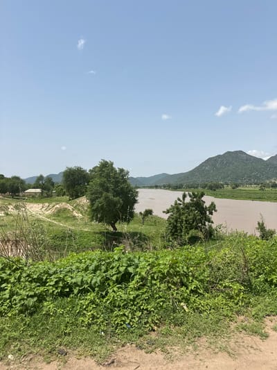The image shows a wide, muddy river flowing through a landscape with green hills in the background and trees and vegetation in the foreground. A building with a light-colored roof is visible on the left bank of the river, surrounded by trees. The sky is blue with a few scattered clouds. The scene appears to be a rural or semi-rural area.