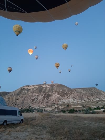 The image depicts a scene at dawn in Cappadocia, near the city of Avanos, Turkey, characterized by numerous hot air balloons ascending into a clear blue sky. In the foreground, a white van is parked on dry, grassy terrain. Behind the van, the distinctive geological formations of Cappadocia, with their eroded rock spires and valleys, rise prominently. Several hot air balloons are visible, some in the distance and others closer to the viewer. One balloon, prominently in the upper left, is partially in the frame and appears to be the viewpoint of the photographer. Another yellow balloon in the upper left quadrant bears the text "Air Kapadokya BALLOON". Other balloons are scattered across the sky at varying altitudes, some illuminated by the rising sun. The overall atmosphere is serene and suggestive of a popular tourist activity.
