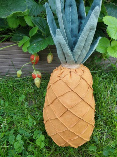 A stuffed pineapple toy with a woven orange exterior and green fabric leaves sits on a patch of green grass, surrounded by clover. In the background, a strawberry plant with several red and green strawberries grows next to a wooden planter. The scene appears to be outdoors, possibly in a garden or backyard. There are no people or animals present. The lighting suggests it is daytime. The image is taken from a slightly elevated angle, looking down at the toy and its surroundings. The overall impression is one of a charming, handcrafted item placed in a natural setting. The location of Chornomors'ke, Ukraine is not discernible from the visual information provided in the image.