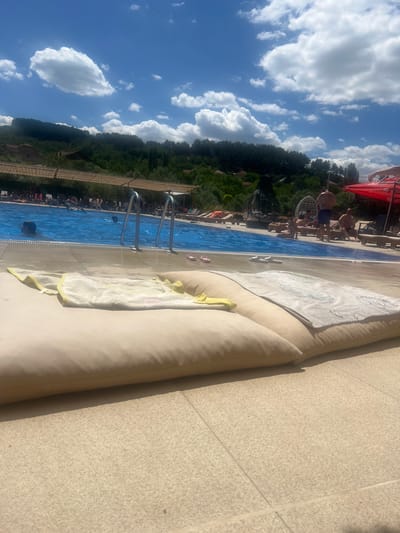 The image depicts a sunny day at an outdoor swimming pool, likely a resort or a public pool, located in Delchevo, North Macedonia. The foreground is dominated by two large, beige lounging cushions on a tiled surface. A white and yellow towel is casually draped over one of the cushions, and another towel with a patterned design lies on the second cushion.
In the background, a vibrant blue swimming pool is visible, with several people enjoying the water. Some are swimming, while others are relaxing by the poolside. The pool area is lined with lounge chairs and umbrellas, and beyond that, a lush, green hillside with scattered buildings and trees forms the backdrop. The sky is a clear blue, dotted with fluffy white clouds, suggesting pleasant weather. The overall atmosphere is one of relaxation and leisure.
