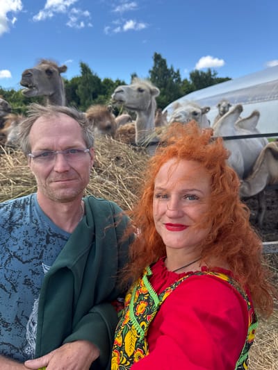 In the city of Tri Ozera, Russia, on a sunny day with a partly cloudy sky, two people are taking a selfie in front of a herd of camels. The man on the left, wearing a blue t-shirt and glasses, has his arm around the woman on the right, who has bright red, curly hair and is wearing a red dress with a colorful, floral pattern. They are standing in a hay-filled enclosure, and several camels are visible in the background, some with their mouths open as if chewing or calling out. The environment appears to be an outdoor enclosure, possibly a farm or zoo, with hay scattered on the ground and a white structure in the background. The overall mood seems cheerful and informal.