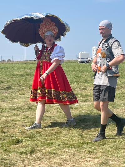 In Surakovo, Russia, on a grassy field under a clear blue sky, a woman wearing a traditional red dress with embroidered floral patterns and a distinctive ornate headdress walks to the left, holding a large, decorative umbrella with a metallic silver interior. She has braids and appears to be mid-stride. Beside her, to her right and walking in the same direction, is a man wearing a grey t-shirt, a grey vest adorned with patches and pins, a grey bandana, and black shorts. He is looking towards the left. In the background, a white trailer or RV is visible, along with some fencing and a few other indistinct figures. The ground is covered in dry, mown grass.