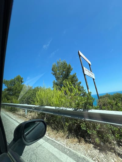 The image shows a roadside view on a clear, sunny day with a bright blue sky. A metal guardrail runs along the side of the road, separating it from a slope covered in dry grass, gravel, and various green shrubs and trees. In the background, a sign with text is mounted on two wooden posts. The upper part of the sign is white with red lettering that appears to say "D. KM. 2." The lower sign is also white with black lettering, reading "La Montagna" and below that, "Trattoria." Beyond the vegetation and the sign, a glimpse of the blue sea can be seen. The foreground includes the interior of a car, with a portion of the car's window and a side mirror visible on the left. The scene appears to be located on a coastal road, potentially near Mattinata, Italy, given the presence of "La Montagna Trattoria," which is a known establishment in the area.