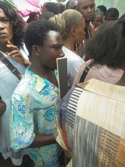 The image displays a dense crowd of individuals in an outdoor setting in Ajah, Nigeria. The people, appearing to be of African descent, are closely packed. In the central foreground, a person with short, dark, curly hair wears a light blue, green, and white patterned short-sleeved shirt and holds a vertical, rectangular object with a black face and a light-colored reverse near their chin, looking to the right. To the left, a woman with dark, wavy hair wears a white dress with a brown shoulder strap; her left hand is raised towards her chin. To the right, the back of a person in a multi-patterned shirt with vertical stripes is visible. The midground and background contain additional individuals, including a woman with a patterned headwrap and a bald man in a striped shirt. Partial views of pink/white and green fabric structures, likely awnings or tents, are discernible in the upper background, indicating a public gathering or market environment.