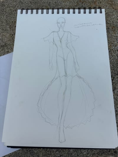 This media file depicts a sketch of a person drawn in pencil on a white spiral-bound sketchbook. The sketch is oriented vertically and shows a full-body figure wearing a voluminous dress. The figure is drawn in a loose, gestural style, with some areas more detailed than others. The head is simply rendered with eyes, nose, and mouth. The torso and arms are outlined, and the dress appears to have a fitted bodice with a very full, tiered skirt that has a jagged, decorative edge.
The sketchbook is placed on a textured surface that appears to be outdoor ground, possibly stone or gravel. There are some faint yellow stains on the paper.
Visible text is written in cursive on the upper left side of the page, appearing to be in Spanish. It reads: "recuallado zig-zag + micro indeable Indumbre". Given the location context provided, "O Casal, Spain," this text might be a note related to the drawing's subject or inspiration.
The time of day and weather are not discernible from the image. The overall impression is of an artistic work in progress, possibly a fashion sketch.