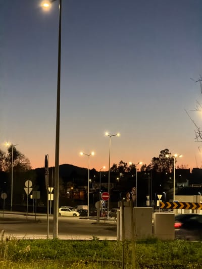 The image depicts an urban street scene at twilight in Penafiel, Portugal. The sky displays a gradient from deep blue overhead to an orange-pink hue at the horizon, indicating dusk or dawn. Numerous streetlights with illuminated, bright white fixtures are active across the landscape.
In the immediate foreground, an area of green grass and sparse vegetation is visible. To the right, two light-colored utility boxes stand near the roadside. The midground features a multi-lane road or intersection with various traffic signs. These include a prominent red circular "No Entry" sign with a white horizontal bar, several white rectangular and triangular informational or directional signs, and a black and yellow chevron hazard marker positioned on the right side of the road.
Multiple vehicles are present on the roadway. A white hatchback, possibly a Volkswagen Golf, is stationary on the left side of the road. Behind it, other dark-colored vehicles are visible. On the right, a dark-colored vehicle is in motion, evidenced by motion blur on its activated red taillights. No people are discernible.
The background consists of forested hills or mountains with scattered residential buildings, some of which display illuminated windows.