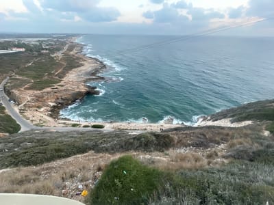The image captures a scenic view of the Mediterranean coastline in the Mate Asher Regional Council, Israel. A winding road snakes along the rocky shoreline, with cars visible on it. The sea is a vibrant turquoise, with waves crashing against the rugged cliffs. Vegetation, a mix of dry grasses and green shrubs, covers the hillside in the foreground, with some scattered litter visible on the ground. In the distance, a city skyline is faintly visible on the horizon. The sky is overcast with scattered clouds, suggesting a possibly cool or breezy day. A few people are seen walking along the coastal path and on the beach area. The overall scene portrays a natural landscape being encroached upon by human development.