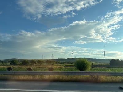 The image depicts a rural landscape with wind turbines in the background, likely located near Urbnisi, Georgia. The foreground features a guardrail separating a road from a field. In the field, there are several dark, rounded objects that appear to be hay bales. The sky is blue with scattered white clouds, suggesting it is daytime. The overall scene is one of agricultural land with renewable energy infrastructure.