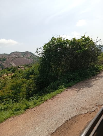 The image depicts a rural landscape with a paved road in the foreground, curving to the right. The road surface shows signs of wear and tear, with patches of exposed earth and gravel. To the left of the road, lush green vegetation grows, including trees and bushes. In the background, a prominent rocky hill with exposed soil and striations dominates the landscape. The sky is bright and partly cloudy, with a few scattered white clouds. The scene appears to be a natural environment, possibly on the outskirts of a town or village.