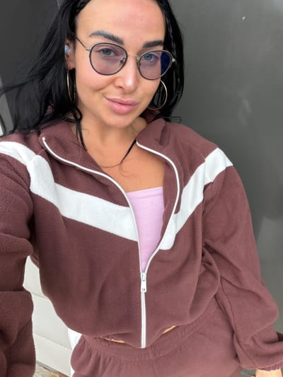 A young woman is posing for a close-up selfie outdoors. She has black hair, fair skin, and is wearing round glasses and gold hoop earrings. She is wearing a brown, fleece zip-up jacket with white stripes over a light pink top. The background appears to be a dark-colored surface, likely a wall or building exterior. The location is not discernible as Sust'e-Konets, Russia from the available image.