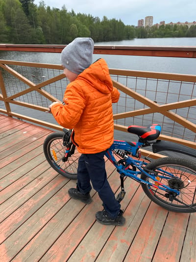 A boy in a gray hat and orange puffer jacket stands on a wooden deck, leaning over a railing, seemingly looking out at a body of water. A blue bicycle is propped against the railing next to him. The location appears to be a waterfront area in Grebnevo, Russia, based on the buildings and natural environment visible in the background. The wooden deck has a grid-like railing and is constructed of planks.