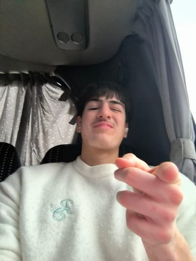 The scene depicts a young male individual seated inside a passenger vehicle in Zlatina, Bulgaria. The male, with dark hair and a light mustache, is positioned centrally in a low-angle selfie. He wears a white, long-sleeved crew-neck sweater featuring a light blue embroidered monogram on the left chest. White wireless earbuds are visible in both ears. His eyes are squinted, and his lips are pursed, suggesting a deliberate expression. His right hand is extended towards the camera, with fingers curled into a pointing gesture. The background shows the interior of the vehicle, including a light gray ceiling with an overhead console containing two circular vents or lights. Dark patterned upholstery is visible on the seatback behind the individual. Drawn curtains flank the subject: a gray curtain with a faint patterned texture is on the left, and a plain dark gray curtain is on the right. Ambient light illuminates the interior.
