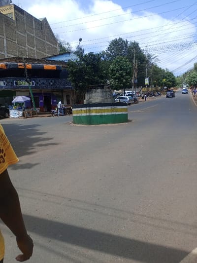 The image depicts a street scene in Murang'a, Kenya, under a partly cloudy sky.

In the immediate foreground, the lower left quadrant shows a dark-skinned arm and shoulder, partially clothed in a yellow t-shirt with a dark graphic resembling paw prints and the letters "LA." Prominent shadows extend across the asphalt road surface from an unseen object or person, indicating direct sunlight.

The midground is dominated by a circular, low-rise concrete street island painted with horizontal bands of green, yellow, and black. A stacked, cylindrical concrete structure is centered on top of this island, from which a thin metal rod or pipe protrudes. Behind the island, several green trees with dense foliage are visible.

To the left of the central island, a row of commercial buildings lines the street. The upper portion of one building is constructed of exposed concrete blocks. Below, storefronts feature awnings; one dark awning has orange horizontal stripes and a sign with "3P" visible. In front of these shops, a vendor's stall with a purple and white striped umbrella is set up, displaying items in buckets. A male individual in a white shirt and dark trousers stands near this stall, appearing to interact with a small three-wheeled vehicle.

The right side of the midground features a paved, two-lane road receding into the distance. A silver sedan/hatchback is parked on the left side of this road, followed by a white SUV and other passenger vehicles, including a blue sedan and another silver sedan, further down the street. Multiple motorcycles are parked along the road's edge. Pedestrians are observed walking on the side of the road. Utility poles, supporting a complex array of overhead wires, line the street, interspersed with trees. A street light with a dark fixture is visible near the central island.

The background shows a continuation of the road and more distant trees under the blue sky with scattered white clouds.