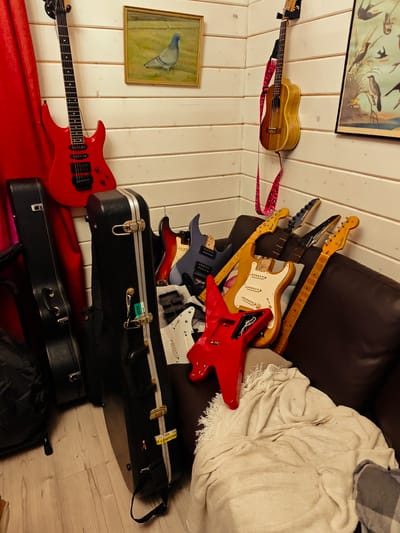 The scene depicts a corner of a room with wood-paneled walls. A red electric guitar leans against the wall next to a red curtain, close to a painting of a pigeon. To the right, a ukulele hangs on the wall near a framed print of birds. Several guitar cases stand on the wooden floor. On a brown leather couch are various disassembled electric guitar bodies and necks. A white blanket is draped over a portion of the couch. The location is specified as Свистуха (кузяевский С/о), Russia. No people are visible.