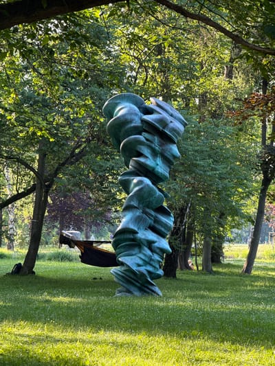A tall, green, abstract sculpture stands in the foreground of a sun-dappled park. The sculpture is made of swirling, layered material, appearing somewhat organic or like stacked stones. Behind it, trees with lush green foliage form a dense canopy. The ground is covered in vibrant green grass dotted with small white flowers. In the mid-ground, between the sculpture and the trees, a dark hammock is suspended between two trees, with a backpack leaning against one of the tree trunks. Further in the background, a few indistinct figures can be seen in the distance, suggesting the park is open to the public. The lighting indicates it is daytime, likely late afternoon or early morning given the long shadows cast by the sun. This scene is located in the Forest Opera park (Opera Leśna) in Sopot, Poland, a popular venue for outdoor performances and a recreational area.